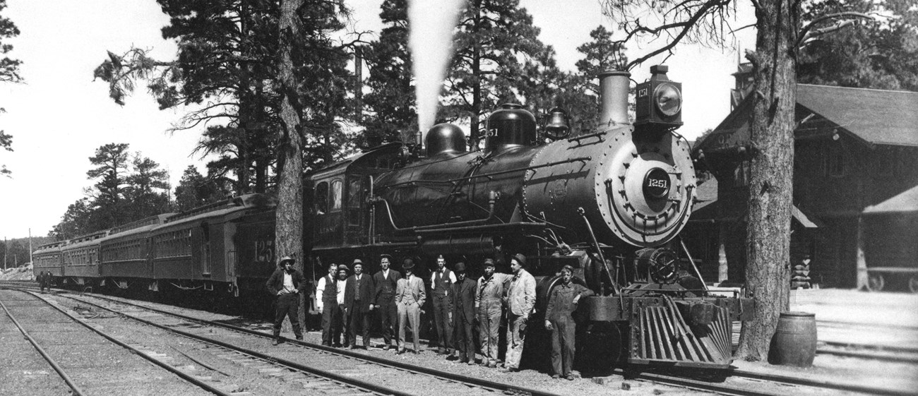 Black and white photo of 11 men standing in a line outside of a steam engine carrying 4 train cars.