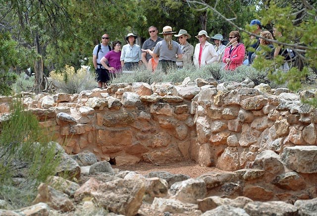 In the foreground, the partial ruin wall, about three feet high. In the background, a ranger has her back to the camera and is addressing a group of 10 people who are taking her tour.