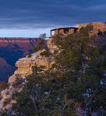 Looking over trees as the ast rays of pinkish sunset falling on rustic stone Geology Museum on the edge of Grand Canyon - seen in the distance