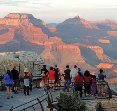 Vermilion sunset light on buttes and peaks within Grand Canyon. A number of people are watching the sunset from behind a metal guardrail