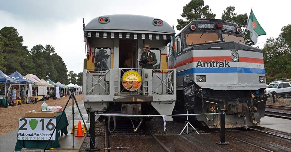 Two trains parked at the Grand Canyon Depot with tents on the left side. In bottom left is a sign reading "2016 National Park Service Centennial." Two trains parked at the Grand Canyon Depot with tents on the left side. In bottom left is a sign reading "2016 National Park Service Centennial."