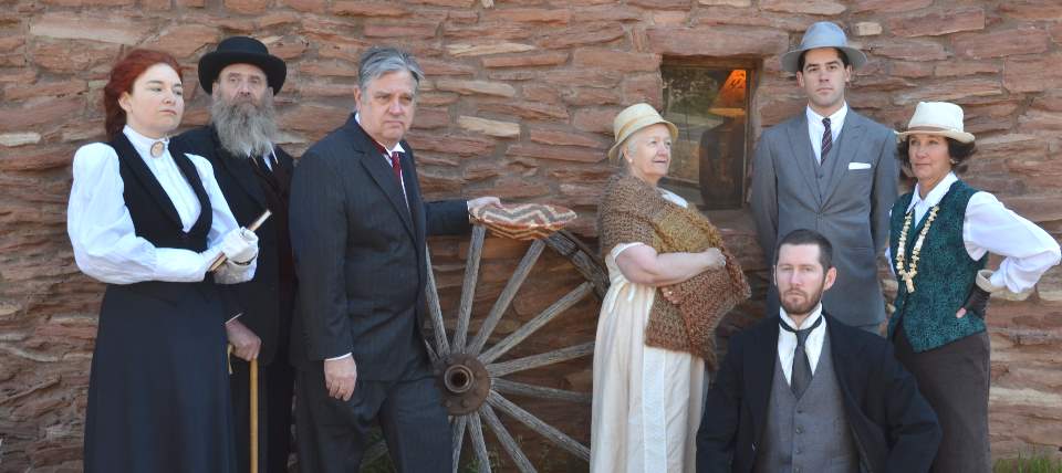 Wearing turn of the 19th Century clothing, 7 members of the Echoes From the Canyon living history production are posing in front of an adobe wall. A large wooden wagon wheel is in the center of the photo.