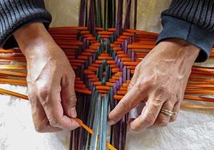Two hands weaving a colorful basket of orange and purple yucca leaves with diamond shaped designs.