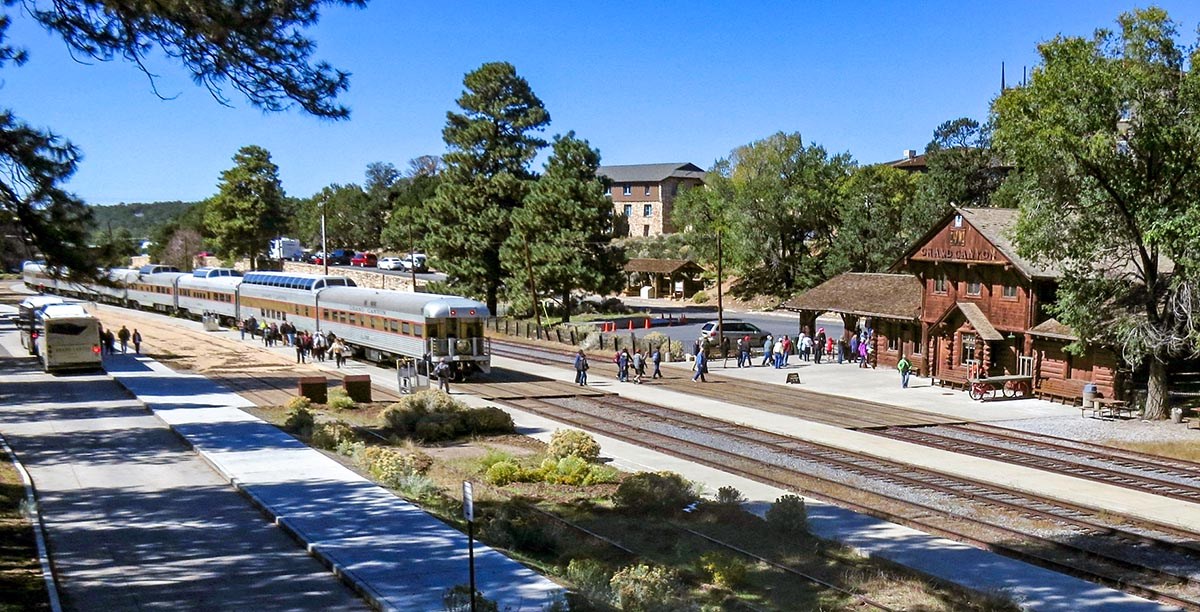 overview of a passenger train that has arrived at a two story log railroad depot. Passengers are getting off the train