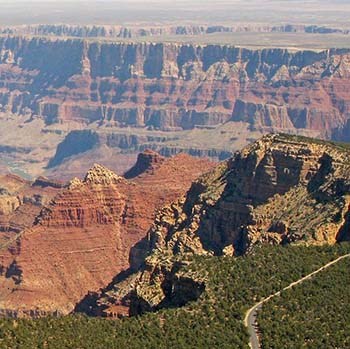 a tree-covered plateau in the foreground, on the right a two-lane highway through the forest and along the canyon's rim. The view opens up into a view of canyon peaks and formations, with the Colorado River below steep cliffs in the distance.