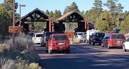 a dozen cars are waiting in two lines at the park entrance station with toll booth lanes covered by rustic style and open-sided roofs.