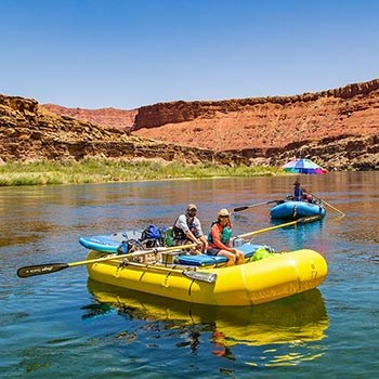 two people wearing life jackets are rowing a yellow inflatable raft on calm river water. a second blue raft is following them.