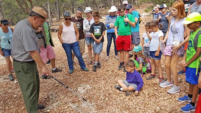 A park ranger pointing a hiking pole at a fossil embedded in the rock beneath his feet, with a crowd of people watching around him.