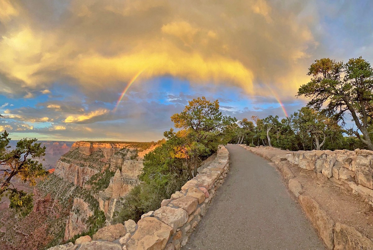 A paved footpath along the sheer edge of a canyon appears to be underneath the arch of a rainbow below a mass of yellow clouds at sunset.