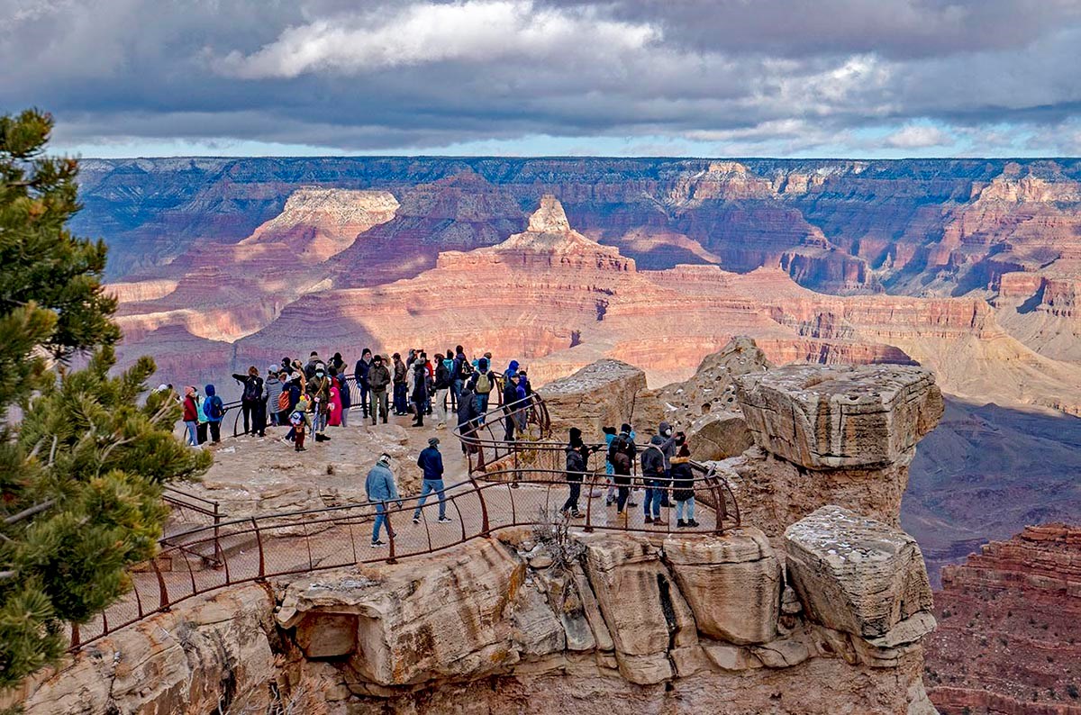 Around 30 sightseers behind railings at a scenic overlook with colorful peaks and cliffs in the background and a blanket of clouds overhead.