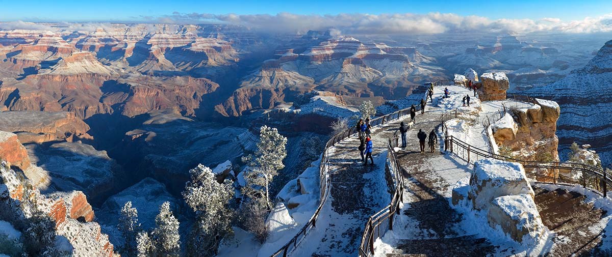 a few people behind railings at a scenic overlook viewing a vast canyon landscape of peaks and cliffs with bands of snow accenting colorful rock layers.