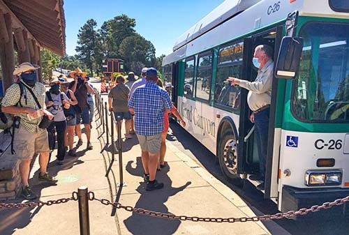 A group of park visitors wearing summer clothes and hats are in line and boarding a green and white bus. The driver standing in the front door is counting them as they are boarding.