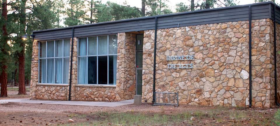 A modern, rectangular stone building with two large windows on the left side, and a door in the middle. The right side is all stone with metal letters that spell Shrine of the Ages.