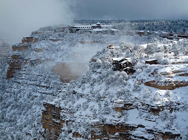 several buildings and a large rustic hotel on the edge of a canyon cliff, with a thick layer of snow covering everything as fog is clearing.