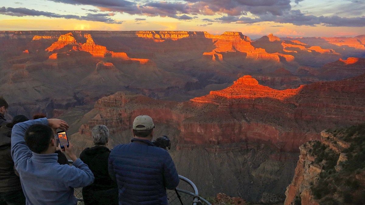Several sightseers behind a guardrail at a scenic overlook are taking photos of a glowing red sunset light illuminating the tops of cliffs and peaks, within a vast canyon landscape.