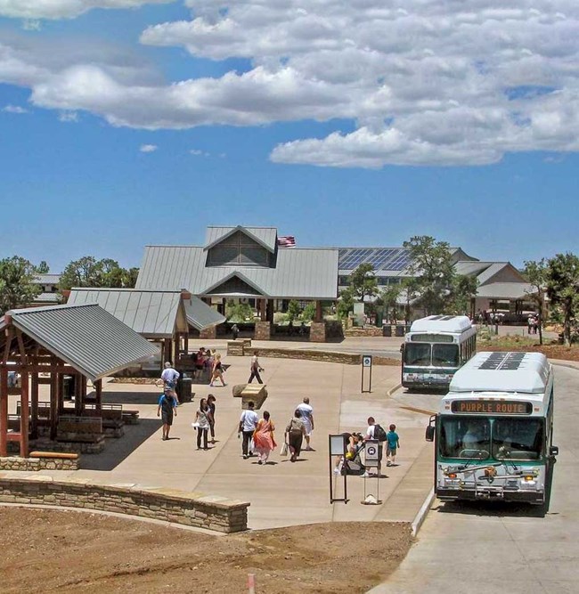 People standing near two white and green buses at an outdoor bus terminal with several shade structures and informational signs.