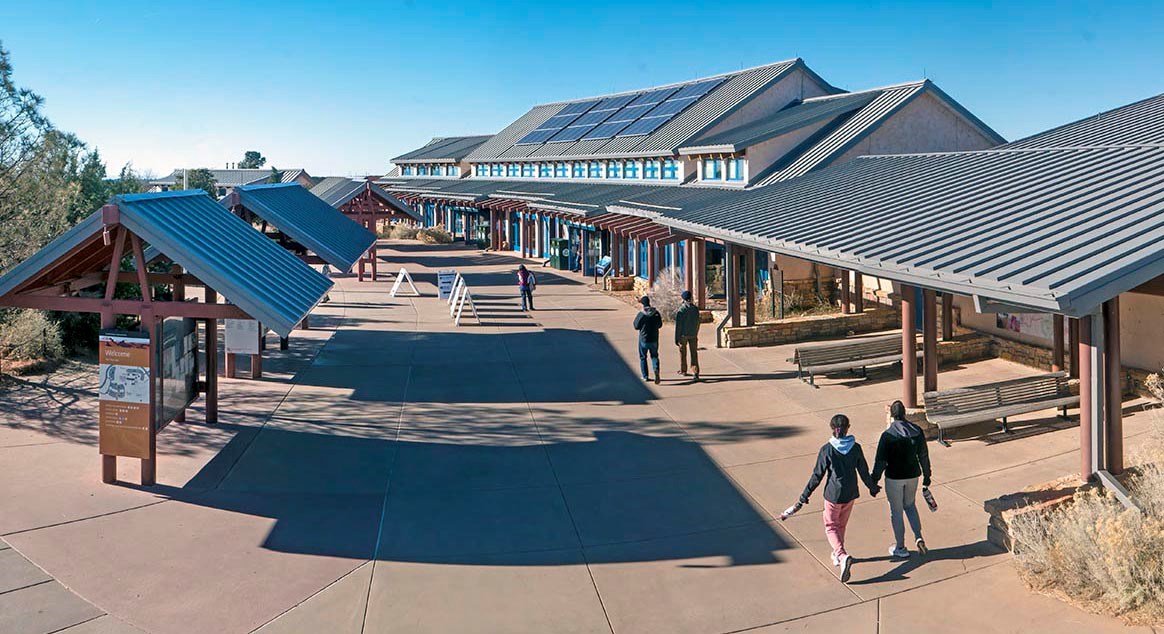 view down and across a concrete plaza with a long narrow building with a gabled roof on the right. several people are walking towards the building.