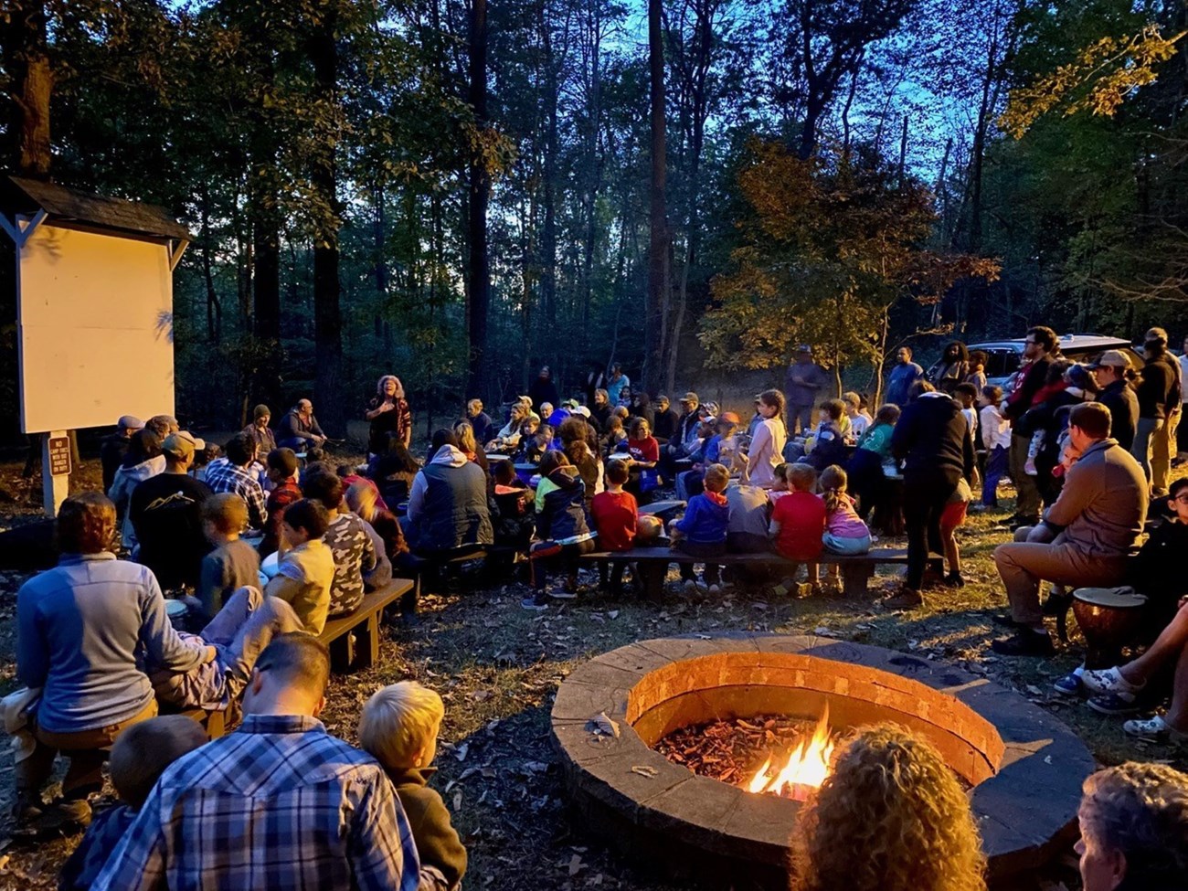 one hundred people sitting on benches enjoying a talk with a campfire in the forefront