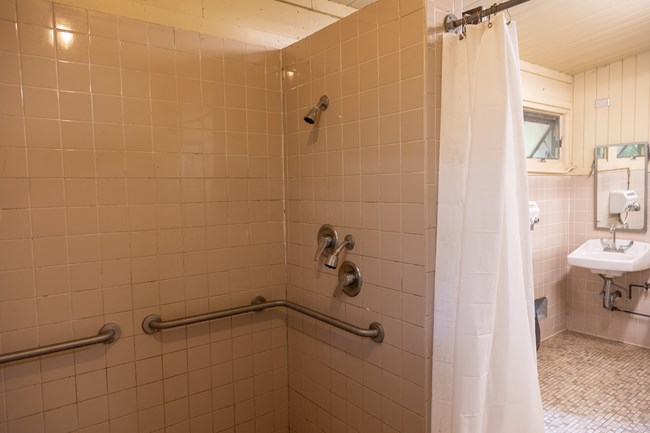 The inside of a shower with tan colored tiles, two grab bars, two fixed shower heads, and two water on/off handles