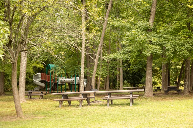 Concrete picnic tables sit adjacent. In the background is a playground with slides, rope, and tubes connecting everything together.