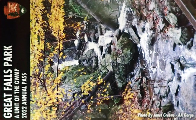 Great Falls Park A Unit of the GWMP Annual Pass 2022, photo by Janet Greene AA Gorge, shows a small rocky waterfall bordered by small trees with leaves in fall yellow colors.
