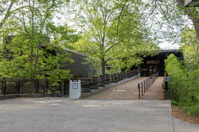 A wide paved ramp with handrails leads towards the second story of a brown building surrounded by trees.