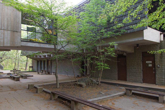 A paved area with benches, tables, and landscaping next to a two-story building with an enclosed bridge arching over the courtyard.