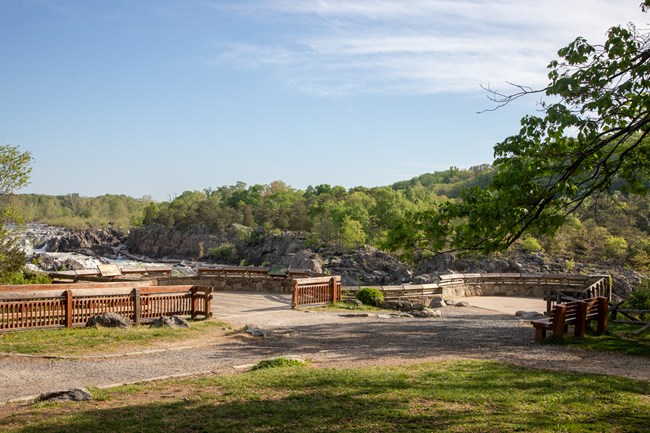 A gravel path leading to a wooden and stone viewing platform overlooking a river lined with trees.