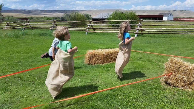 Two children running a sack race through hay bale obstacle course.