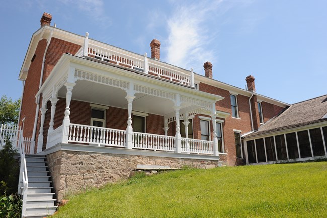 Blue sky above, looking up at three story home. Brick three story home with ornate covered porch painted white on 2nd story and white wooden sided section of house to right.