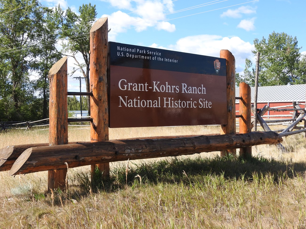 Large sign, NPS black band on top, arrowhead symbol on right, "Grant-Kohrs Ranch National Historic Site" in white text on brown background