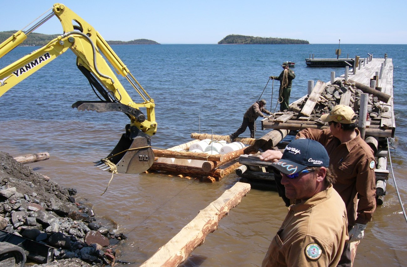 Four people and equipment at a dock construction site at the edge of a lake and island on the horizon.