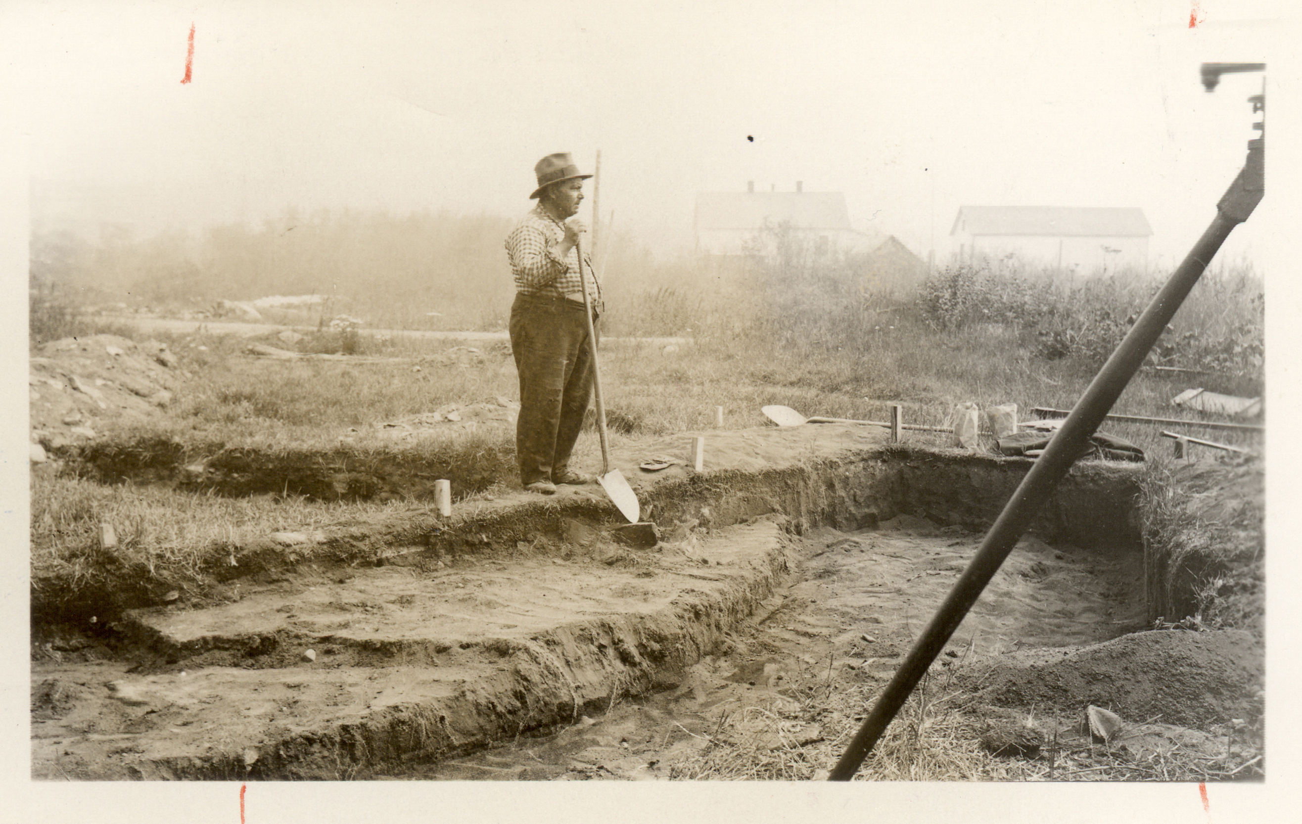 Black and white photo of a person holding a shovel in a terraced dirt area with markers.