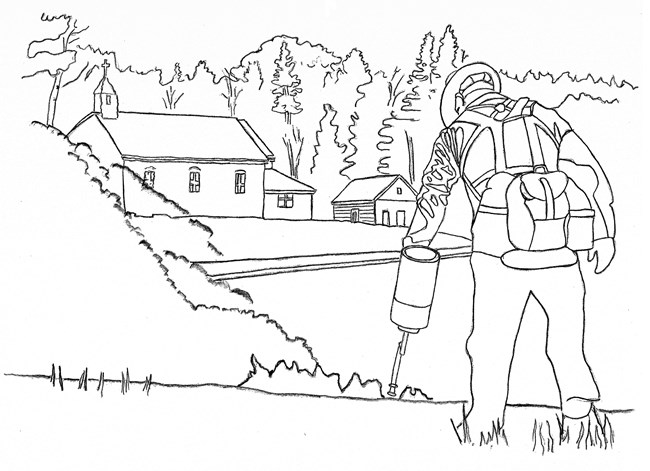A person wearing fire protective gear, holding a drip torch in front of a fire and distant buildings.