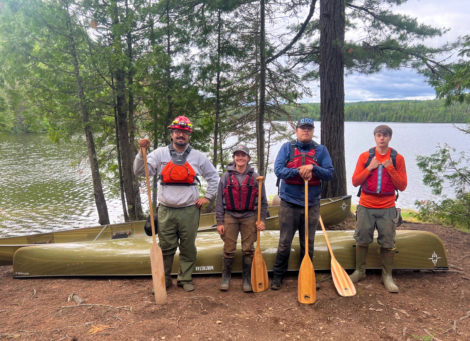 Four people wearing life vests and holding paddles in front of a two Kevlar canoes at the edge of a lake.