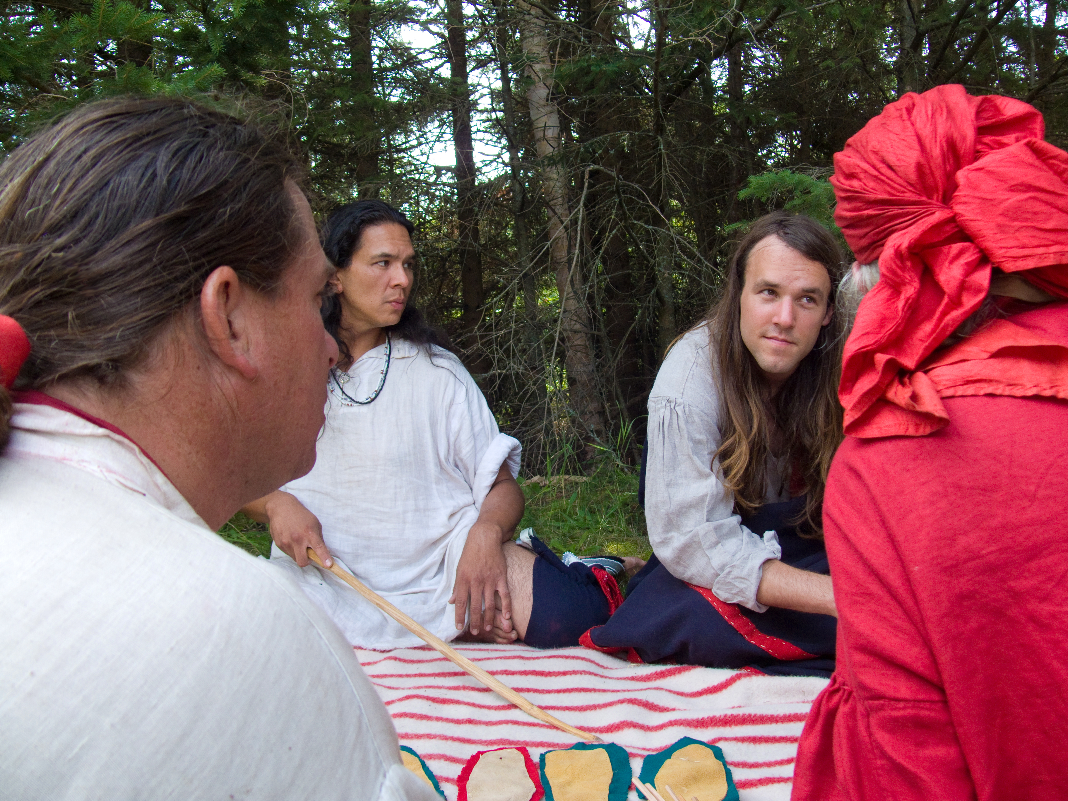 Four people in historic clothing facing each other around a striped blanket with leather pieces on it.