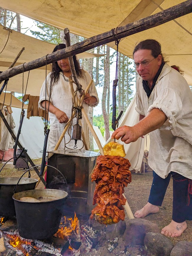 A canvas shelter with two people in historic clothing, cooking over a fire.