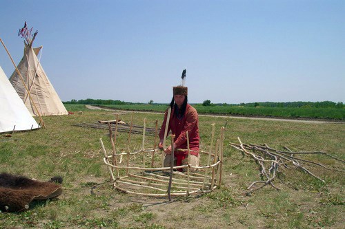 A man in native dress tying sticks into the shape of a boat in a grassy field with canvas tipis beside him.