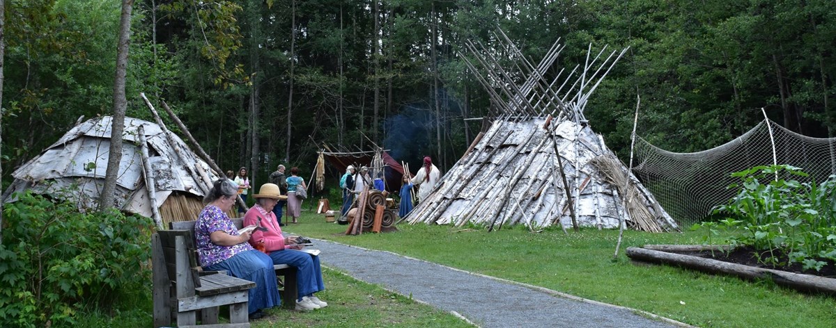 Modern facsimile of a traditional Ojibwe village with visitors resting on a bench in the foreground.