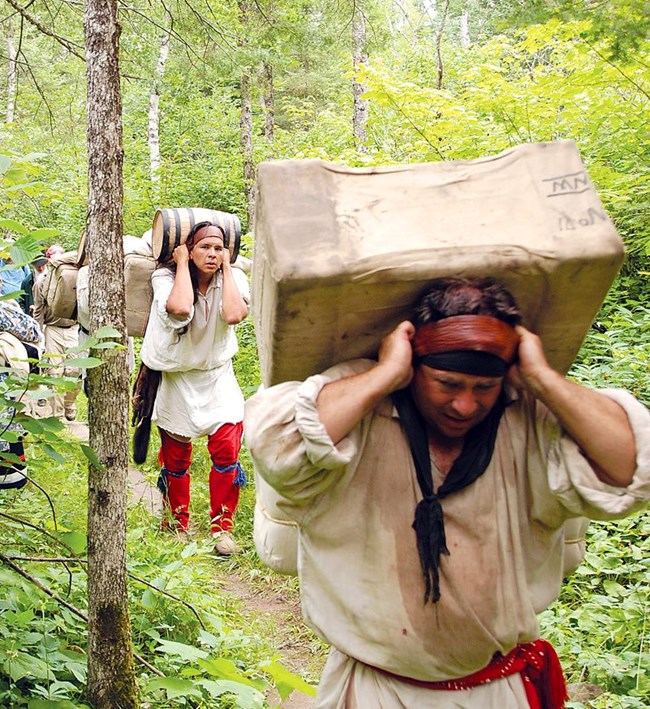 People dressed in historic voyageur clothing on a trail through the forest, carrying heavy bales strapped to their heads.