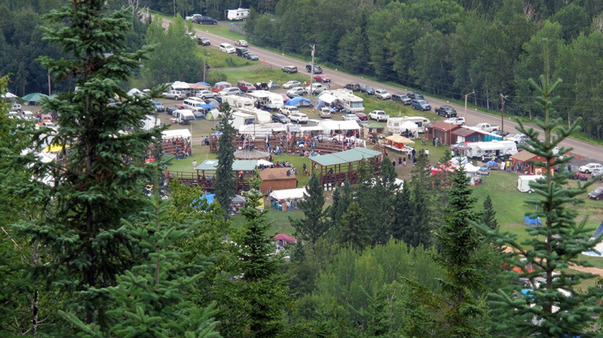Looking down from a hillside at a powwow in progress with many people in colorful clothing, dancing and watching, bleachers, and a line of vehicles along a roadway/