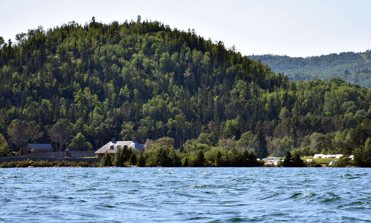 A stockade and buildings at the base of a hill, viewed from the water.