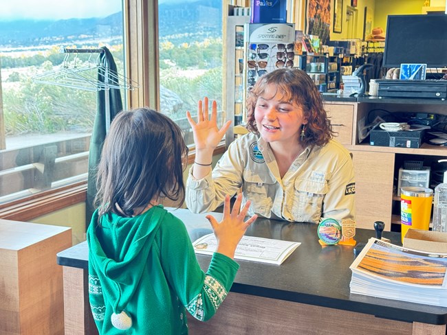 A Conservation Legacy Individual Placement Intern Holds their right hand up in front of a young child during a junior ranger oath ceremony at the park information desk