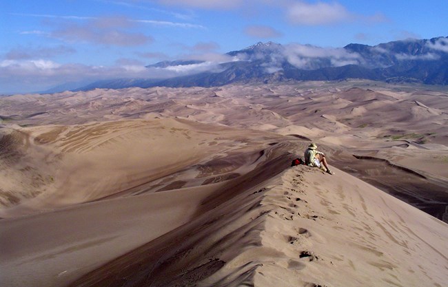 Person sitting on ridge of sand dune with dunefield and mountains in background