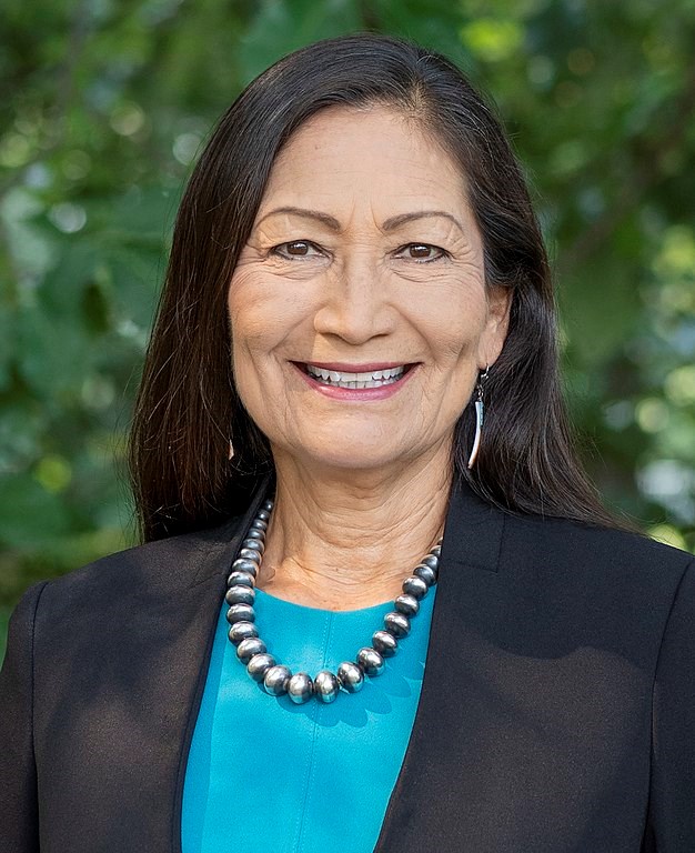 Woman wearing a blazer, blouse, and beaded necklace smiling into the camera with green trees in the background