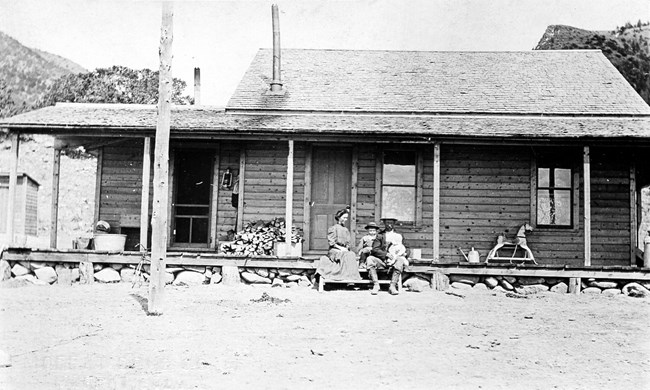 A family sits on the porch of a large cabin at the base of the mountains.