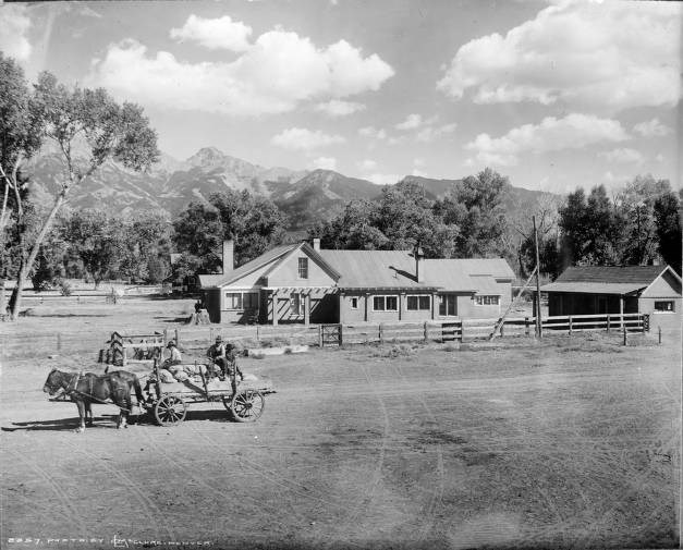 Historical black and white photo of a ranch at the base of mountains, with a wagon and people in the foreground