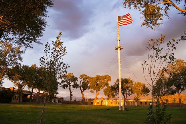 Fort Garland's garrison flag flies over the grassy parade ground, with adobe buildings in a square around it at sunset