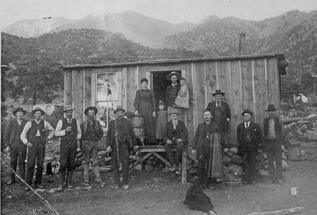 In this historical black and white photo, a family stands on the porch of a cabin surrounded by many miners.