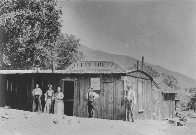 Historical black and white photo of a small hotel with women and men standing outside. In the background are mountains.